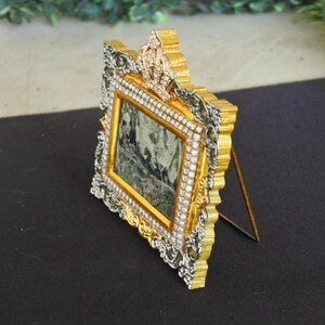 Op de afbeelding: Een rechthoekige fotolijst met een versierde goud- en zilverkleurige rand met ingelegde doorzichtige strass-steentjes. De bovenkant van de lijst is versierd met een kroonmotief dat is bezet met strass-steentjes. Een dunne metalen standaard.