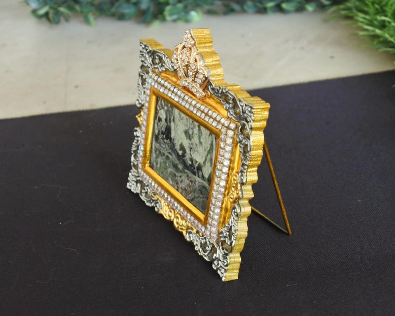Op de afbeelding: Een rechthoekige fotolijst met een versierde goud- en zilverkleurige rand met ingelegde doorzichtige strass-steentjes. De bovenkant van de lijst is versierd met een kroonmotief dat is bezet met strass-steentjes. Een dunne metalen standaard.
