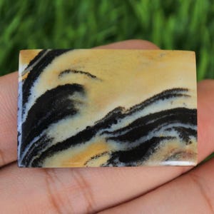 May include: A rectangular gemstone with a swirling pattern of black and cream colours. The polished stone is held in a hand, showcasing its natural beauty and unique design. The stone's surface has a glossy finish.