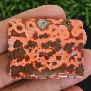 Puede incluir: Una piedra preciosa rectangular con un patrón único. La piedra presenta un color base naranja vibrante con patrones circulares e irregulares de color marrón oscuro y rojo. Una pequeña inclusión de color claro es visible en la parte superior.