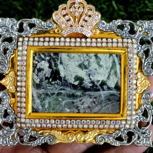 May include: A rectangular frame featuring a central green and white marbled stone inlay. The frame is decorated with gold and silver filigree, a crown-shaped top, and a border of small, iridescent rhinestones surrounding the stone centre.