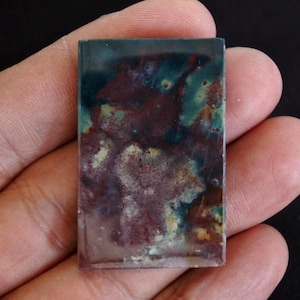 May include: A rectangular gemstone with a unique pattern of colors. The top portion is a deep teal, transitioning to a mix of burgundy, cream, and yellow hues. The stone is held in a hand, showcasing its polished surface and natural beauty.