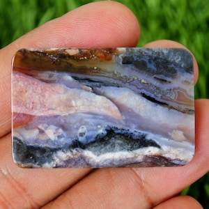 May include: A rectangular gemstone with rounded corners, displaying a natural pattern of colours. The stone features shades of pink, white, blue, black, and brown, creating a unique, abstract design. The polished surface reflects light, highlighting the stone's natural beauty.