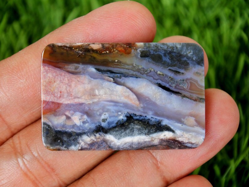 Pode incluir: Uma pedra preciosa retangular com cantos arredondados, exibindo um padr&atilde;o natural de cores. A pedra apresenta tons de rosa, branco, azul, preto e castanho, criando um design abstrato &uacute;nico. A superf&iacute;cie polida reflete a luz.