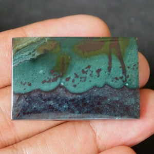 May include: A green, blue, and brown rectangular cabochon gemstone with a layered pattern. The gemstone has a smooth, polished surface.