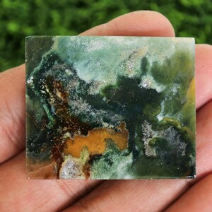 Peut inclure: Une pierre pr&eacute;cieuse carr&eacute;e et polie avec un motif abstrait naturel. La pierre pr&eacute;sente un m&eacute;lange de teintes vertes, brunes et jaunes, cr&eacute;ant un aspect marbr&eacute; unique. La pierre est tenue dans une main sur un fond vert flou.