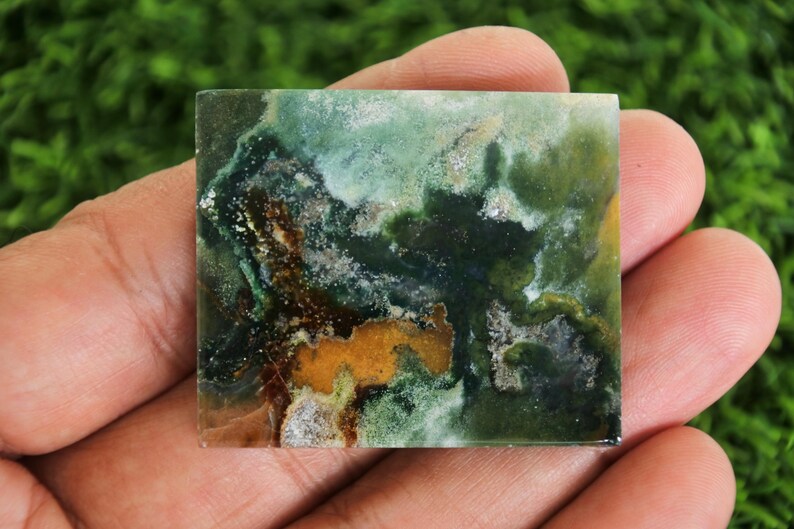 Peut inclure: Une pierre pr&eacute;cieuse carr&eacute;e et polie avec un motif abstrait naturel. La pierre pr&eacute;sente un m&eacute;lange de teintes vertes, brunes et jaunes, cr&eacute;ant un aspect marbr&eacute; unique. La pierre est tenue dans une main sur un fond vert flou.