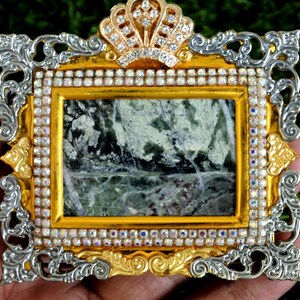 Op de afbeelding: Een rechthoekige broche met een centrale inleg van groen en wit gemarmerde steen. Het frame is goudkleurig met zilverkleurige, sierlijke krulranden, een kroonvormige bovenkant en een rand van kleine, heldere, iriserende strass-steentjes.