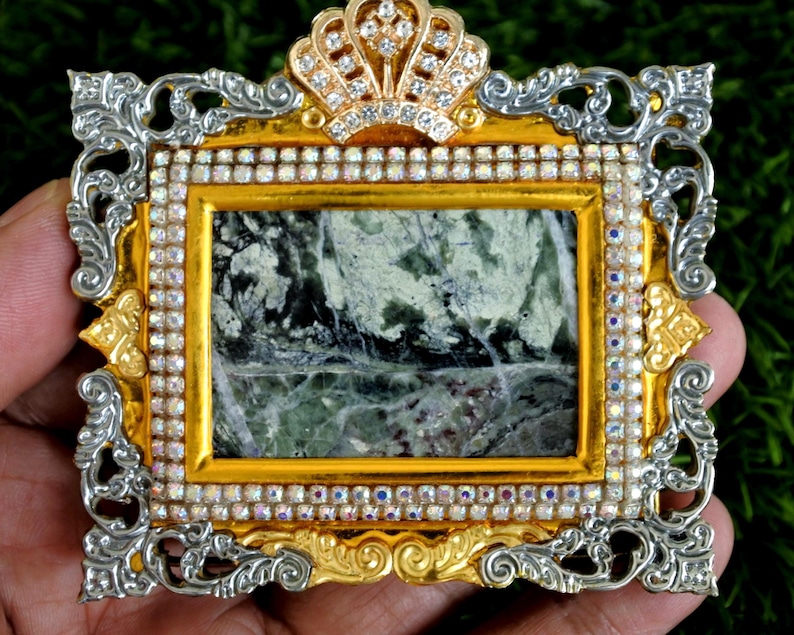 Op de afbeelding: Een rechthoekige broche met een centrale inleg van groen en wit gemarmerde steen. Het frame is goudkleurig met zilverkleurige, sierlijke krulranden, een kroonvormige bovenkant en een rand van kleine, heldere, iriserende strass-steentjes.
