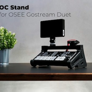 Könnte beinhalten: Ein schwarzer Ständer für den OSSE GoStream Duet Video-Switcher. Der Ständer verfügt über einen integrierten Monitor und ein Bedienfeld mit Knöpfen und Drehreglern. Der Ständer steht auf einem dunklen Holztisch.