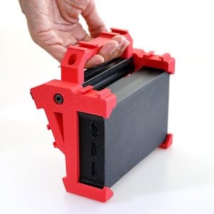 Può includere: Una custodia di trasporto in plastica rossa con maniglia per un piccolo dispositivo elettronico. La custodia ha una superficie testurizzata e un dispositivo nero all'interno.