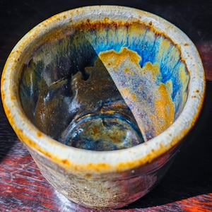May include: A handmade ceramic bowl with a unique glaze. The interior features a blend of blue, gold, and brown hues, while the rim has a cracked, off-white and brown finish. The bowl's design showcases a blend of artistry and functionality.