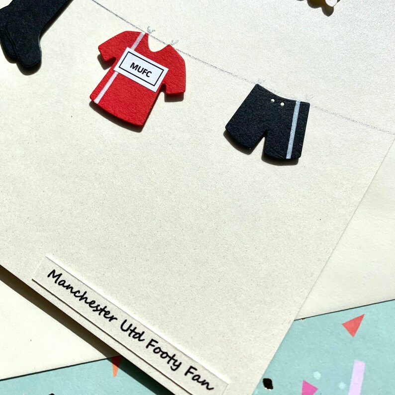 May include: A card with a red and white soccer jersey, black shorts, and the text "Manchester Utd Footy Fan".