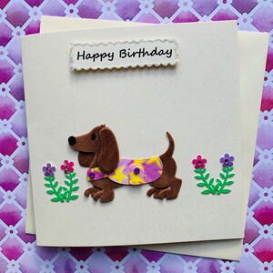 May include: A handmade birthday card featuring a brown dachshund wearing a purple and yellow patterned shirt. The card has a white background with the words "Happy Birthday" in black lettering.