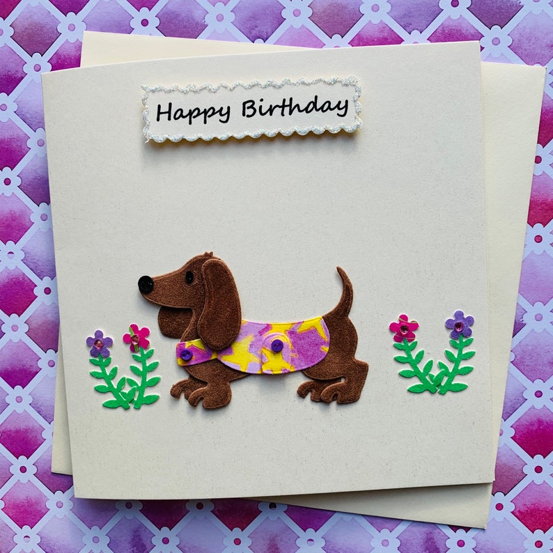 May include: A handmade birthday card featuring a brown dachshund wearing a purple and yellow patterned shirt. The card has a white background with the words "Happy Birthday" in black lettering.