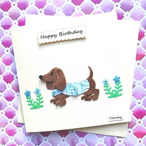 May include: A handmade birthday card featuring a brown dachshund wearing a blue and white patterned shirt. The card has the words "Happy Birthday" and the name "Tommy" written on it.
