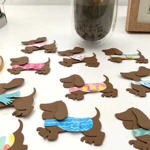 May include: A collection of brown paper dachshund cutouts with different coloured patterned sweaters and scarves. The cutouts are arranged on a white surface.