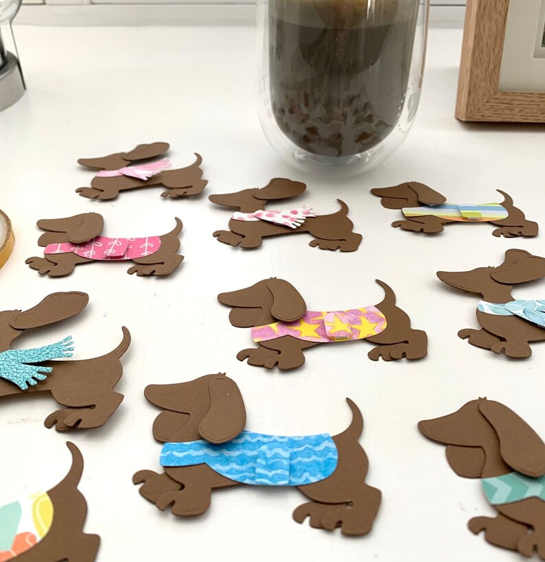 May include: A collection of brown paper dachshund cutouts with different coloured patterned sweaters and scarves. The cutouts are arranged on a white surface.