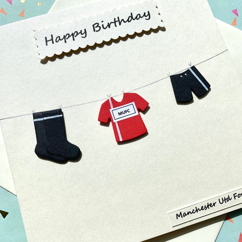 May include: A white card with the words "Happy Birthday" in black script. A red soccer jersey with the letters "MUFC" on the front is hanging on a string with black socks and black shorts. The text "Manchester Utd For" is at the bottom of the card.