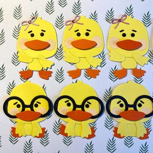 May include: Six yellow duck cutouts with orange beaks and feet. Three ducks have pink bows, and three wear black-rimmed glasses. The ducks are arranged on a white background with a green leaf pattern.