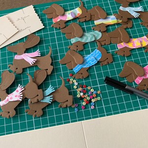 May include: A green cutting mat with brown paper dachshund cutouts. The dachshunds are wearing different coloured scarves and have various patterns. There are also colourful buttons scattered on the mat.