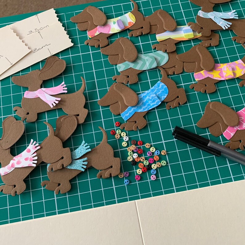 May include: A green cutting mat with brown paper dachshund cutouts. The dachshunds are wearing different coloured scarves and have various patterns. There are also colourful buttons scattered on the mat.
