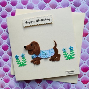 May include: A handmade birthday card featuring a brown dachshund wearing a blue and white patterned shirt. The card has the words "Happy Birthday" and "Tommy" written on it.
