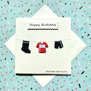 May include: A white card with the words "Happy Birthday" and "Manchester Utd Footy Fan" printed on it. The card has a red and white soccer jersey, black shorts, and black socks hanging on a clothesline.