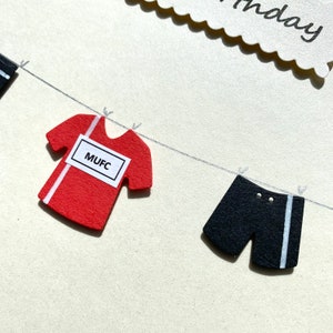 May include: A red and white soccer jersey with the text "MUFC" on a white label hanging on a clothesline with black and white shorts.