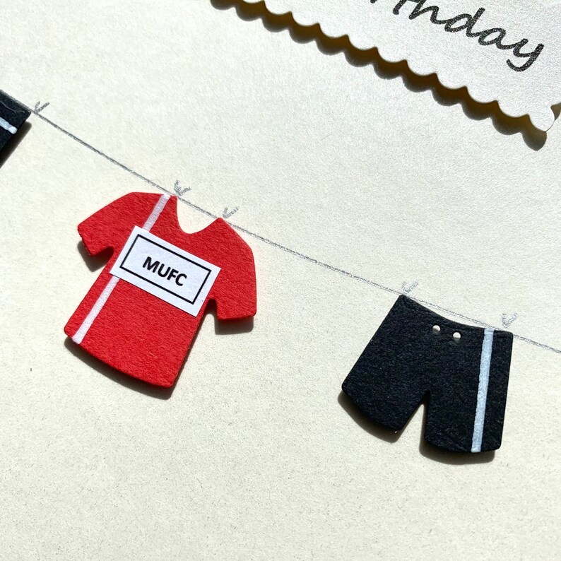 May include: A red and white soccer jersey with the text "MUFC" on a white label hanging on a clothesline with black and white shorts.