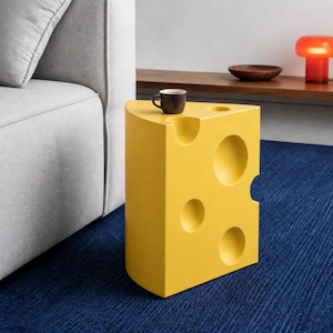Op de afbeelding: Een felgele bijzettafel in de vorm van een punt Zwitserse kaas met diverse ronde inkepingen op het boven- en zijoppervlak. Een kleine, donkerbruine keramische koffiemok staat op het platte bovenoppervlak van de gele kaasvormige tafel.