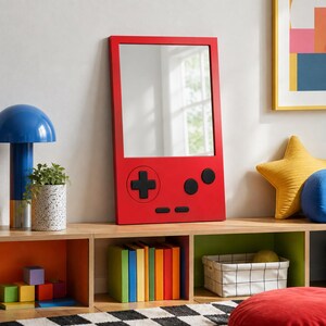 May include: A rectangular wall mirror with a bright red frame designed to resemble a handheld video game console. The mirror features a black directional pad, two circular buttons, and two horizontal buttons on the lower red section.