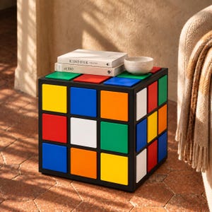Puede incluir: Una mesa auxiliar cuadrada diseñada para parecerse a un cubo de rompecabezas multicolor con un marco negro y paneles individuales rojos, azules, amarillos, naranjas, verdes y blancos. Dos libros titulados KINFOLK TRAVEL y at 100 y un cuenco pequeño están encima.
