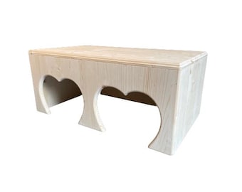 Op de afbeelding: Een houten schuilplaats voor huisdieren met twee boogvormige ingangen. De schuilplaats is gemaakt van natuurlijk hout en heeft een lichtbruine kleur.