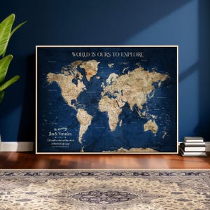 May include: A framed world map with a navy blue background and a beige world map design. The text "WORLD IS OURS TO EXPLORE" is at the top. The names "Jim & Veronica" are below the map. The map is displayed on a wooden floor.