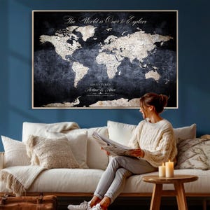 May include: A framed world map with the text "The World is Ours to Explore" and "Adventures Arthur & Alice". The map is in shades of black and white, mounted on a dark blue wall. A woman sits on a white couch, reading.