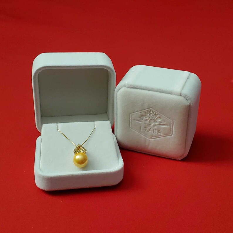 Puede incluir: Una caja de joyer&iacute;a de terciopelo blanco con un collar de perlas doradas en su interior. La caja tiene un logotipo que dice "Izara" en la parte superior.