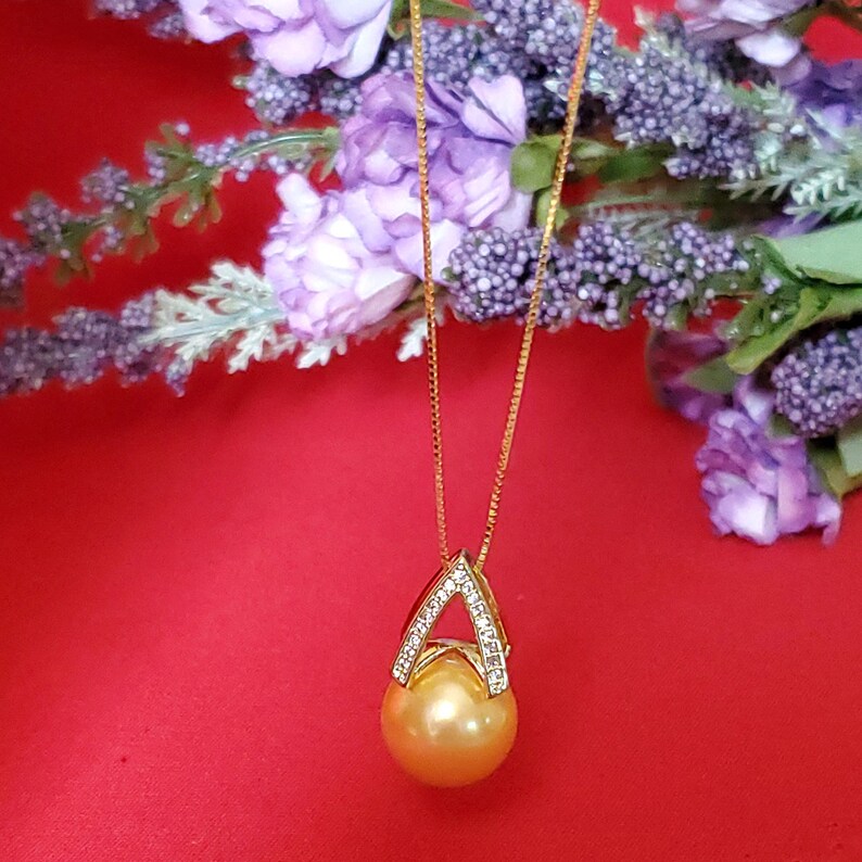 Puede incluir: Un collar de cadena de oro con un colgante dorado que presenta una perla dorada grande, redonda. El colgante est&aacute; acentuado con peque&ntilde;os diamantes transparentes engastados en forma triangular.