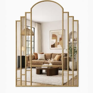 May include: A large wall mirror with a gold-toned metal frame featuring a stepped, arched design. The frame is divided into multiple vertical rectangular sections by thin metal bars, creating a geometric look that reflects a living room interior.
