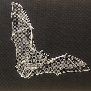 May include: A white embroidered bat on a black fabric background. The bat is in flight with its wings spread out.