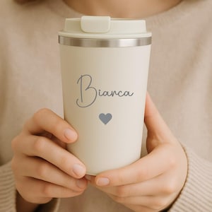 Könnte beinhalten: Cremefarbener Reisebecher mit hellfarbigem Deckel und silbernem Rand. Der Name "Bianca" ist in Schreibschrift geschrieben, darunter ein kleines graues Herz. Der Becher wird von einer Person gehalten.