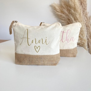Könnte beinhalten: Zwei Kosmetiktaschen aus Canvas mit einem Juteboden. Eine Tasche hat den Namen "Anni" in goldener Schrift mit einem Herz, die andere den Namen "Tha" in rosa Schrift. Beide Taschen haben einen Reißverschluss.