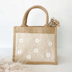 May include: A small, brown burlap tote bag with a white canvas lining. The bag has two handles and features a white daisy pattern on the front.