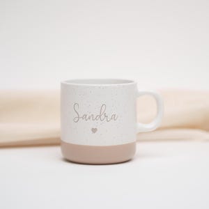 May include: White ceramic mug with a speckled finish and a light pink base. The mug has the name "Sandra" written on it in a cursive font with a small heart symbol below the name.