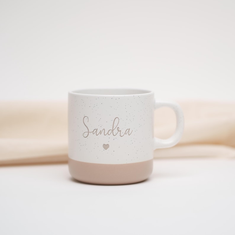 May include: White ceramic mug with a speckled finish and a light pink base. The mug has the name "Sandra" written on it in a cursive font with a small heart symbol below the name.