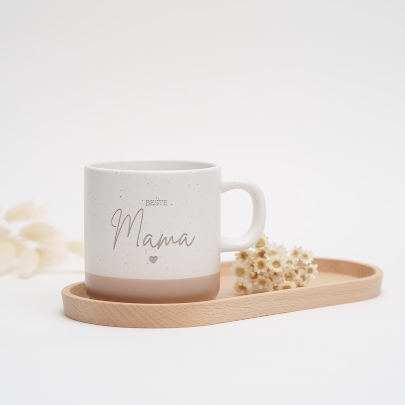 May include: White ceramic mug with a speckled finish and a brown bottom. The mug has the text "Beste Mama" with a small heart symbol printed on it. The mug is sitting on a wooden tray with dried flowers.