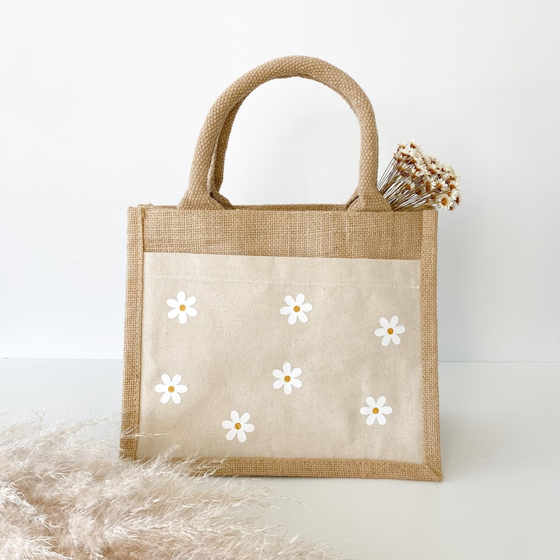 May include: A small, brown burlap tote bag with a white canvas lining. The bag has two handles and features a white daisy pattern on the front.