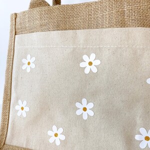 May include: A brown jute tote bag with a white canvas insert. The canvas insert has a pattern of white daisies with yellow centers.
