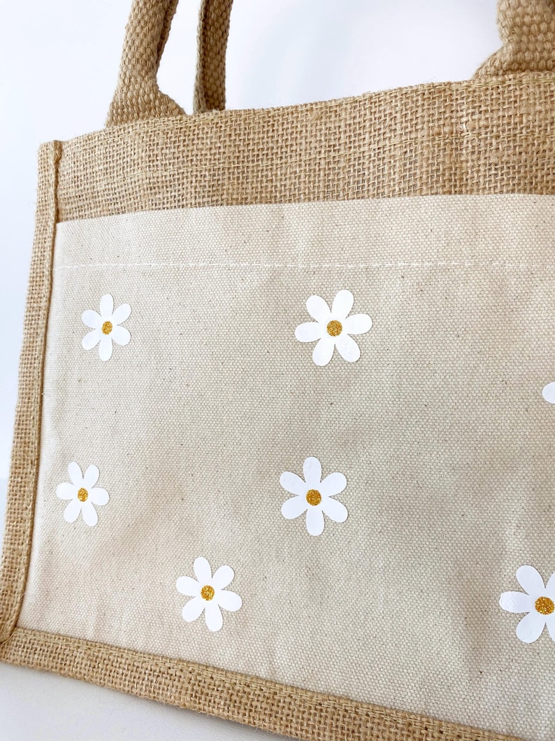 May include: A brown jute tote bag with a white canvas insert. The canvas insert has a pattern of white daisies with yellow centers.