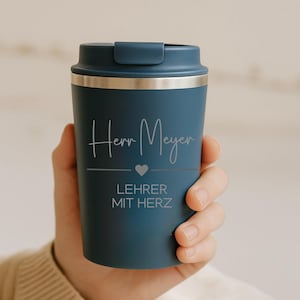 Könnte beinhalten: Ein marineblauer Reisebecher mit silbernem Rand und Deckel. Auf dem Becher steht der Name "Herr Meyer" in Schreibschrift, ein Herzsymbol und die Worte "LEHRER MIT HERZ". Der Becher wird in einer Hand gehalten.